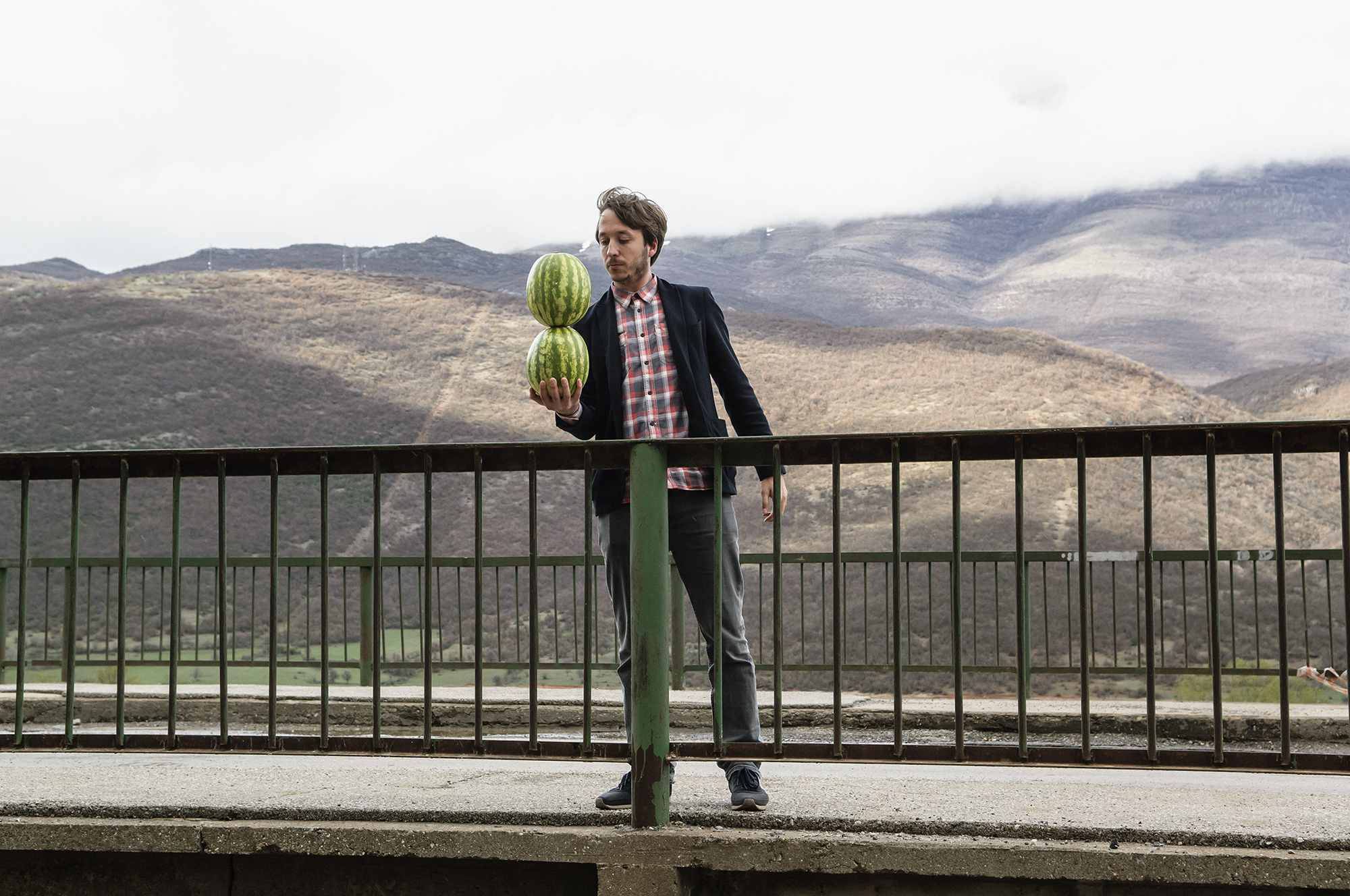 A photograph of a man stood on a bridge in front of hills in sunlight balancing two watermelons in his hand - Driton SELMANI - They say you can't hold two watermelons in one hand