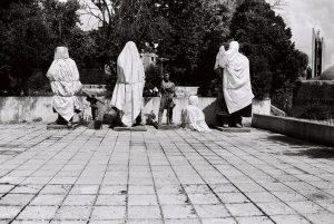 A black and white photograph of three statues wrapped in white sheets on a tiled square