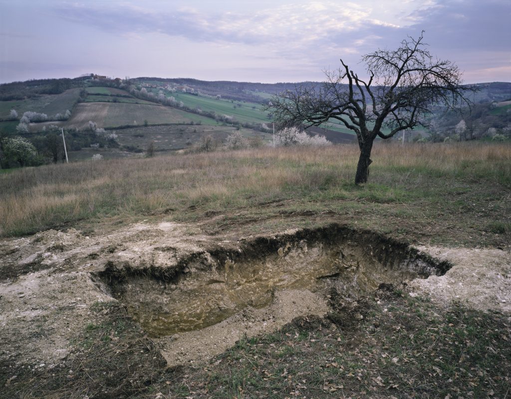 Photograph of a shallow hole in the ground, in a rural landscape with hills in the background and a tree