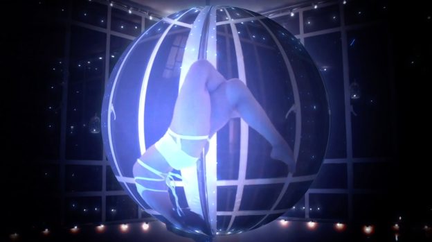 A still from the artist's work displaying a woman pole dancing. The image has a distorted view as you see the woman in a fisheye lens. In the room, there are reflective windows which reflect small circular lights which are dotted around the top of the room. The woman legs are toward the top of the pole crossed over and her head is toward the bottom of the pole. She is wearing a white strappy bralette and white shorts.