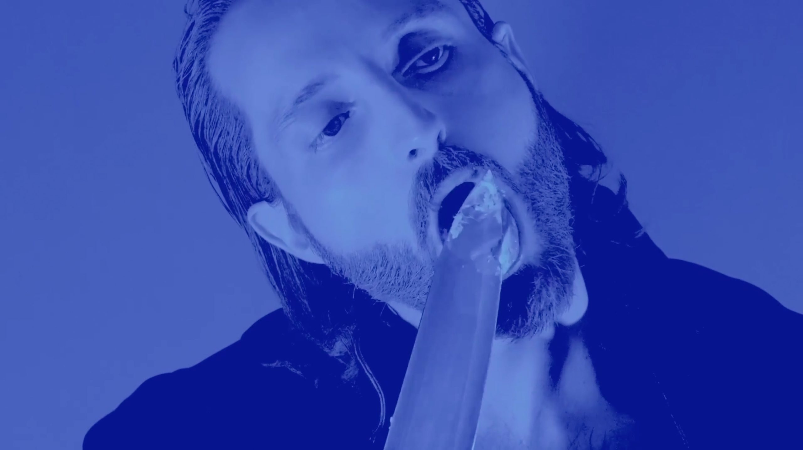 In blue light a white man with beard and swept back hair licks a large knife