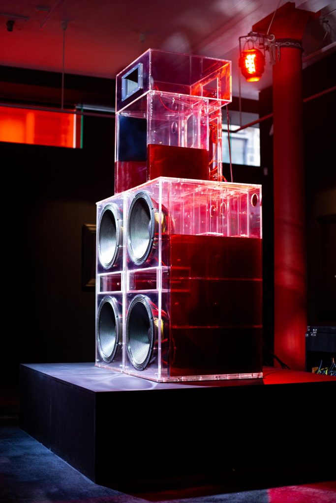 The image is a side view of a sound system as part of the artists work.  The sound system is made up of two speakers side by side and one in the centre and on top. The speakers are translucent with a red tint due to the lighting of the room and they are placed on a small stage. The sound system is in a gallery where you can see two windows at the back, one on the left of the speakers, which is rectangular and has a red tint, and one which is square and transparent and is on the right of the speaker. Also in the room is a pillar behind the system and a red light.